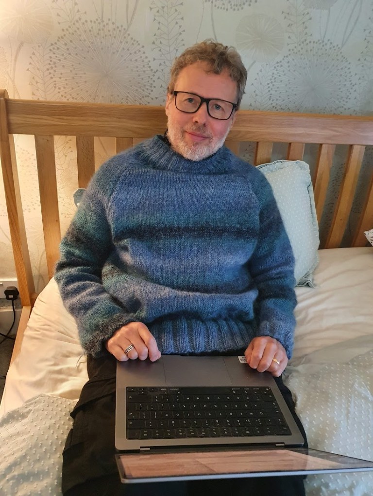Ben sitting in bed looking at the camera and wearing his blue knitted jumper.