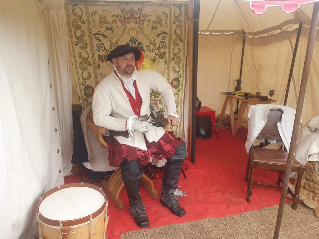 Earl of Mar sitting in his tent with a tapestry hanging behind him.