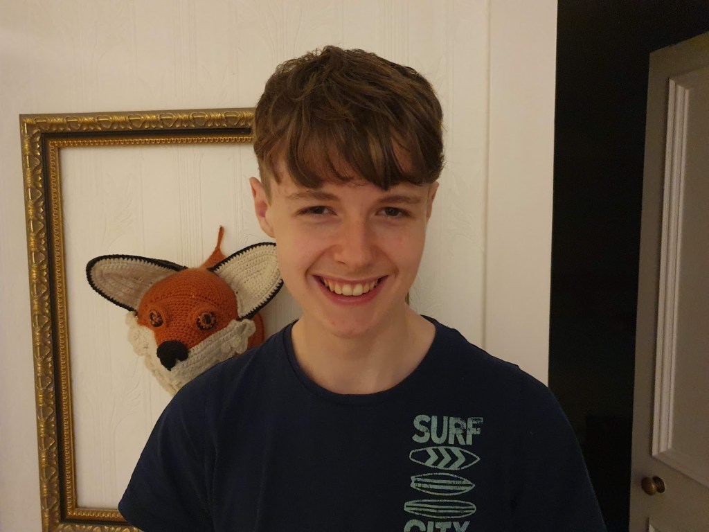 Daniel smiling at the camera. He's wearing a blue t-shirt and my crochet fox head is in the background.