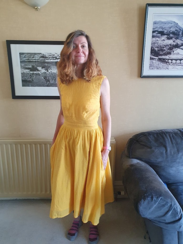 Rachel wearing a yellow dress that has a full circle skirt, is fitted at the top and has button detailing on the back - view from the front