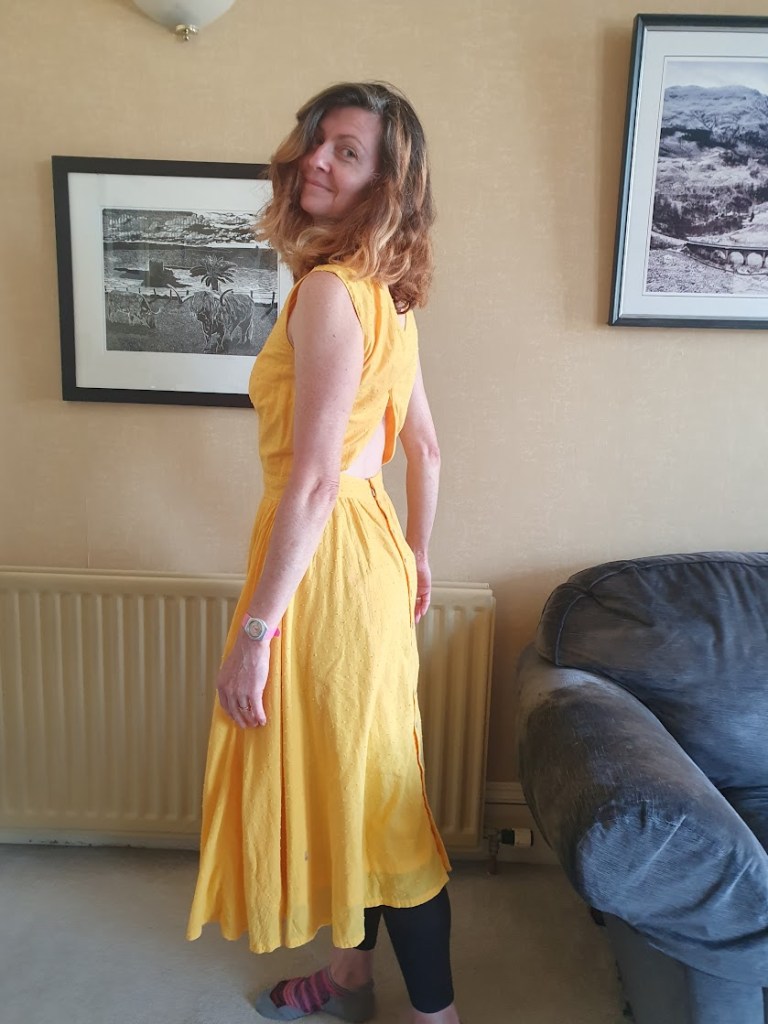 Rachel wearing a yellow dress that has a full circle skirt, is fitted at the top and has button detailing on the back - view from the back