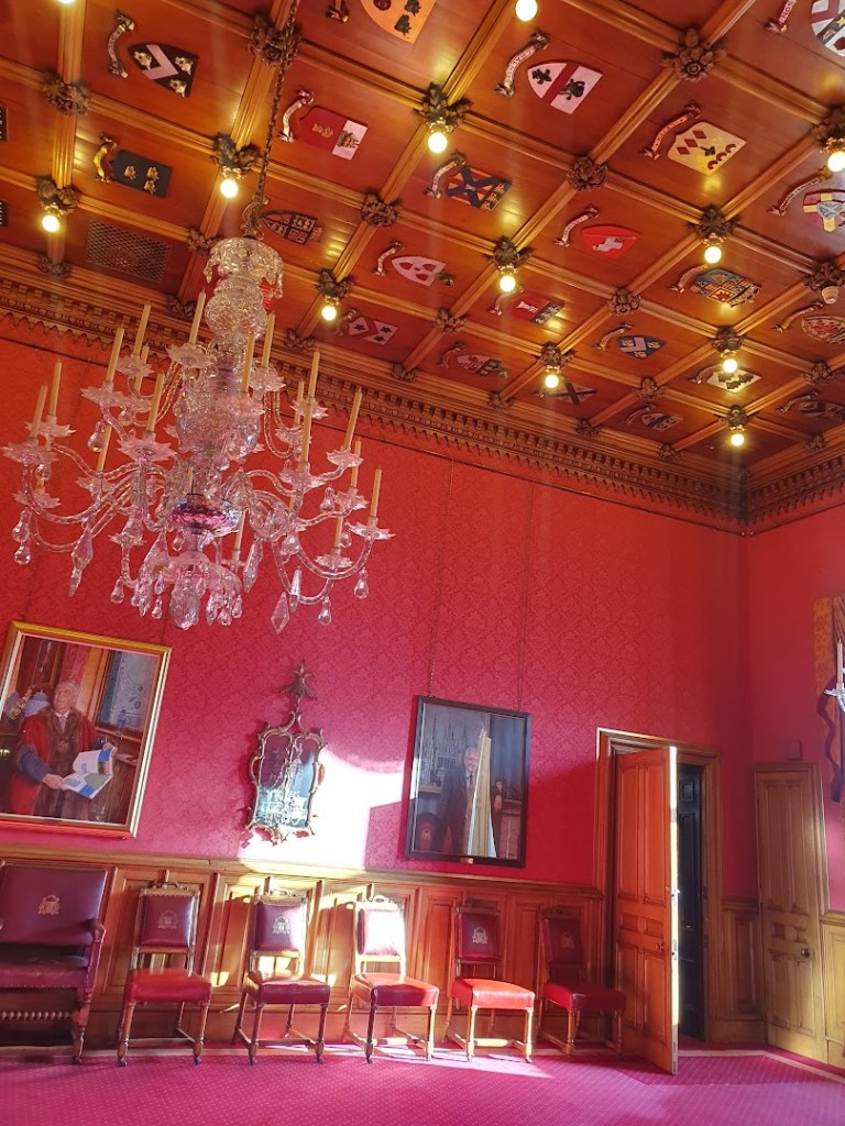 Another photos of a glass chandelier in a grand room with detailing on the ceiling and red walls adorned with paintings of previous provosts. 