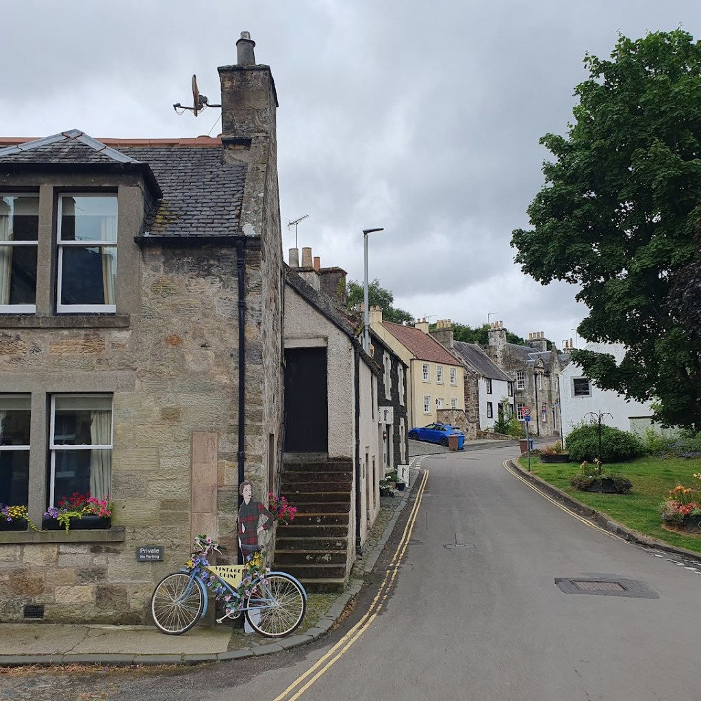 Looking up the street with old buildings on the left and a green on the right.