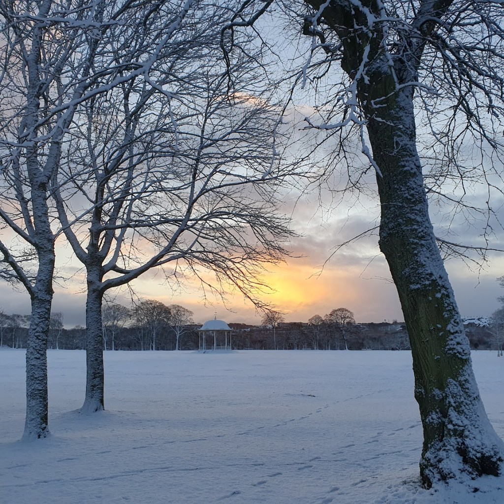 Duthie Park in the snow with the rotunda in the distance and a low rising sun behind giving the sky an orange glow.