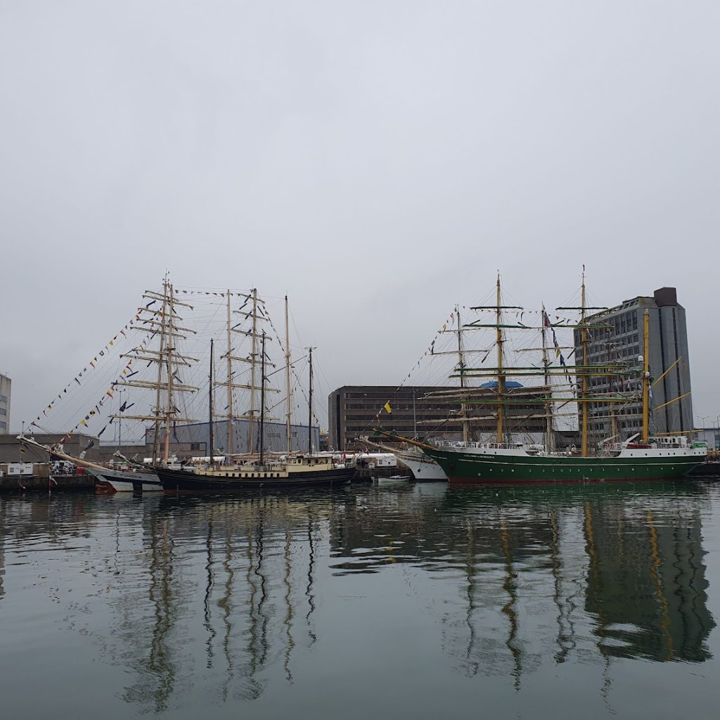 Several tall ships from a distance with some ugly Aberdeen buildings in the background.