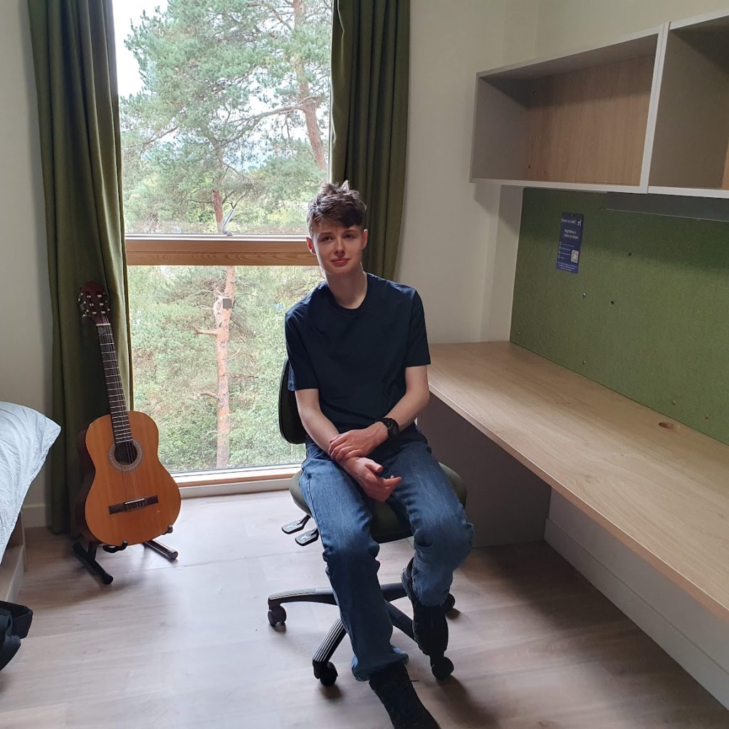 Daniel at his desk in his room with a window and lots of trees behind him. His guitar is next to the bed.