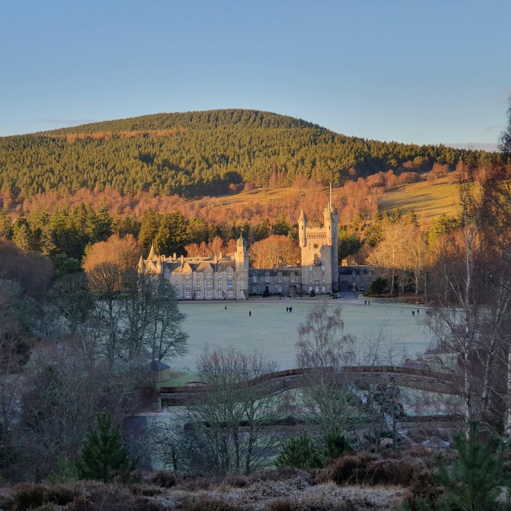 Balmoral Castle from the hill opposite. The sun is shining on it and the hill behind is purple, red and green.