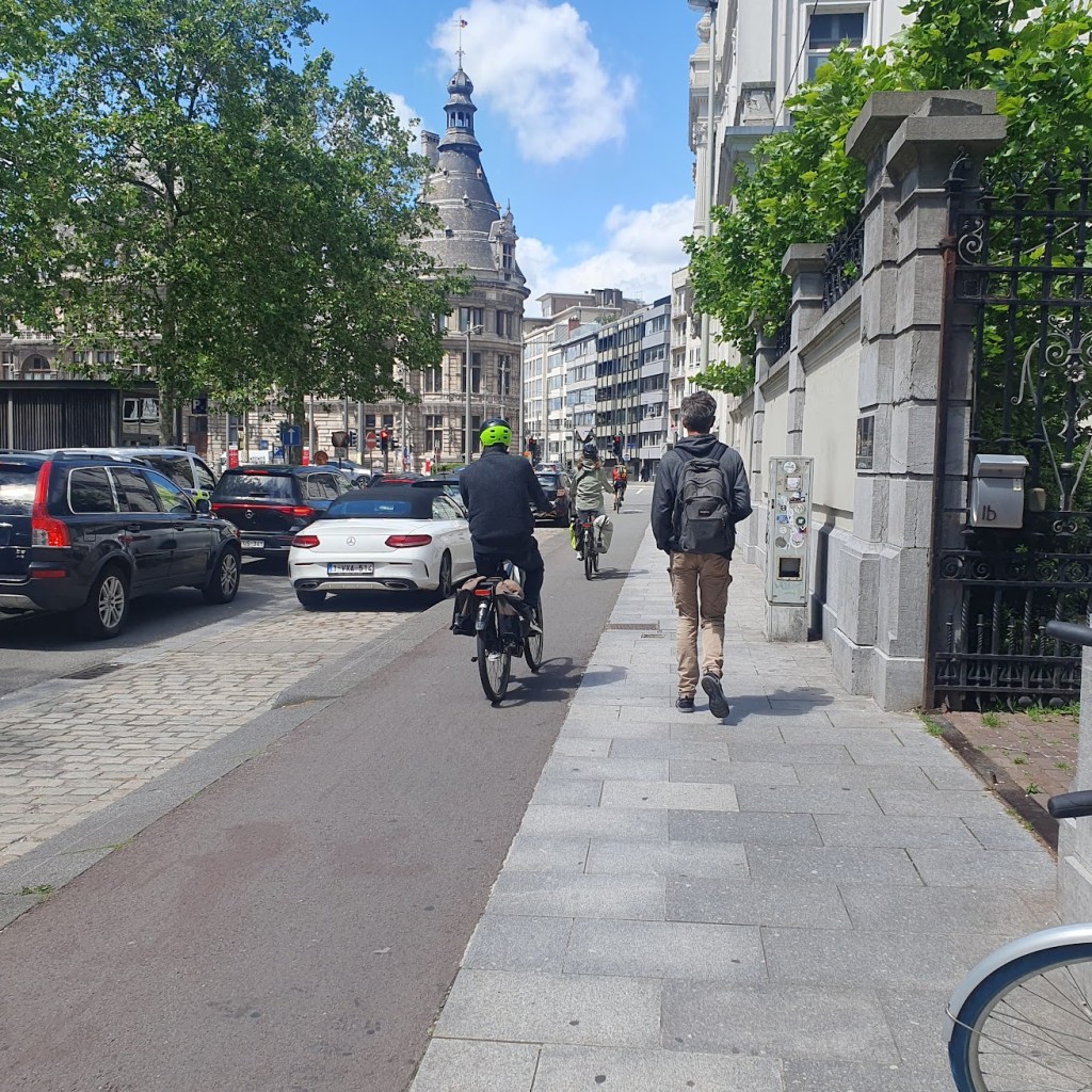 Three cyclists on a segregated bike path between the pavement and parking spaces on the road.