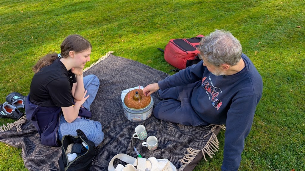 Elizabeth and Ben on the picnic rug with cake and two mugs plus a small bottle of whisky.