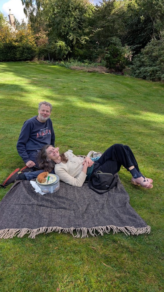 A grassy lawn with Ben and Rachel on a picnic rug plus half an eaten cake.